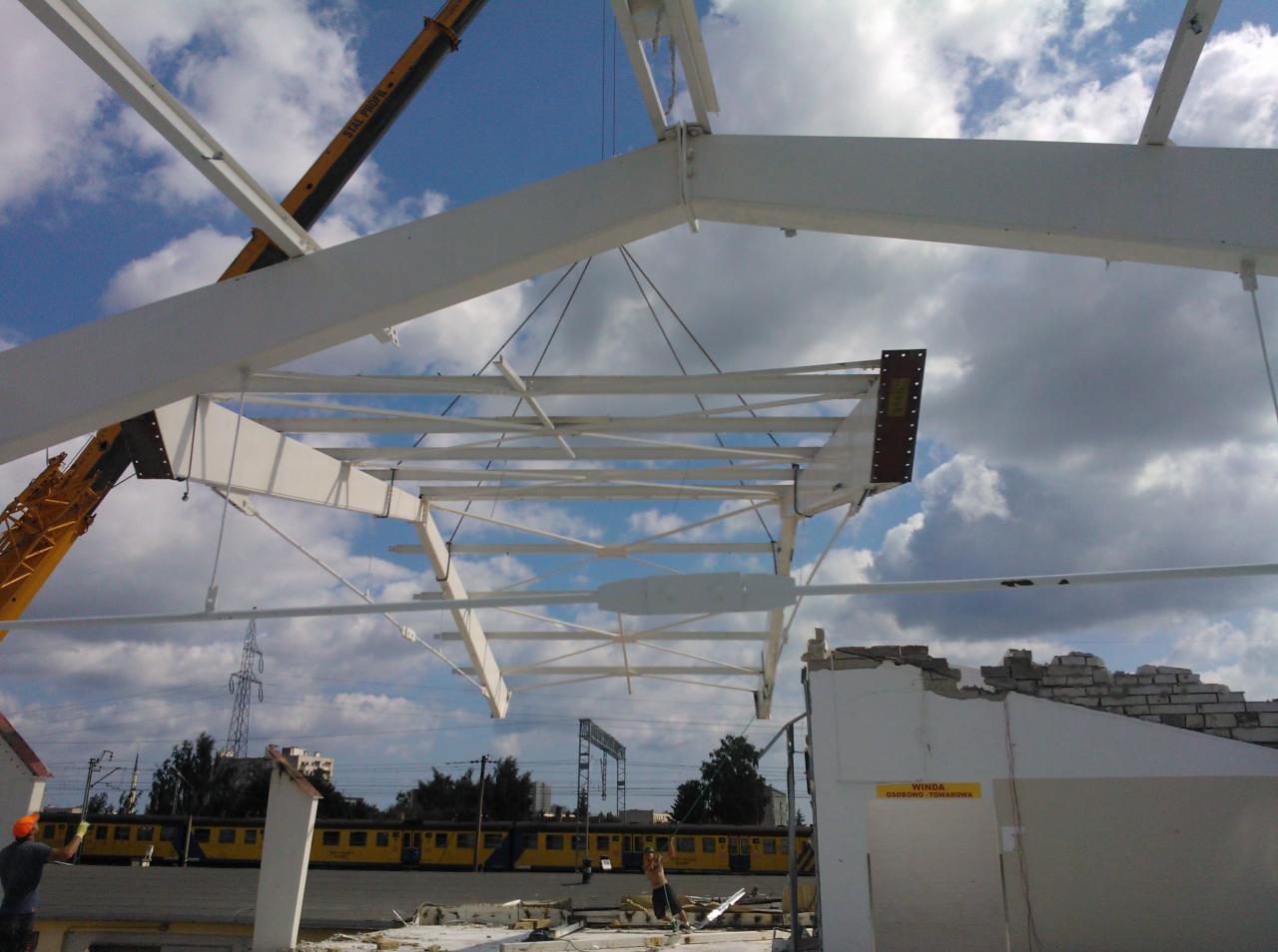 Demontaż białej konstrukcji stalowej z użyciem dźwigu na tle błękitnego nieba i częściowo zburzonego budynku. Widoczny fragment pociągu i pracownicy w kaskach.