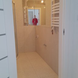 MARCIN GRULKOWSKI - Wykończona łazienka z beżowymi płytkami lastryko na podłodze i ścianach, widoczna kabina prysznicowa i grzejnik drabinkowy. W lustrze odbija się osoba w czerwonej bluzie.