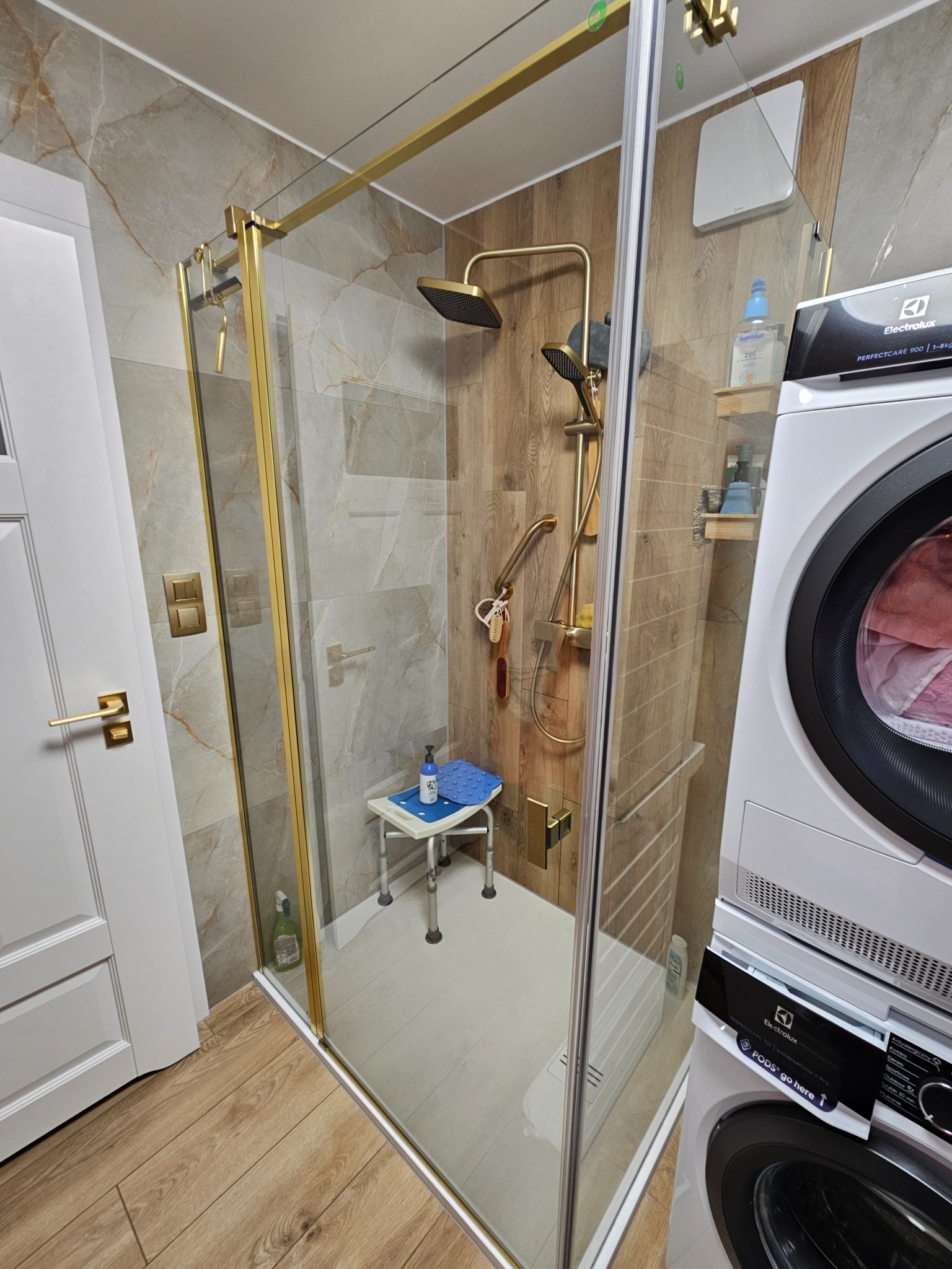 Nowoczesna łazienka z kabiną prysznicową typu walk-in, wykończoną złotymi profilami. W tle pralka i suszarka Electrolux. Na podłodze drewnopodobne płytki.