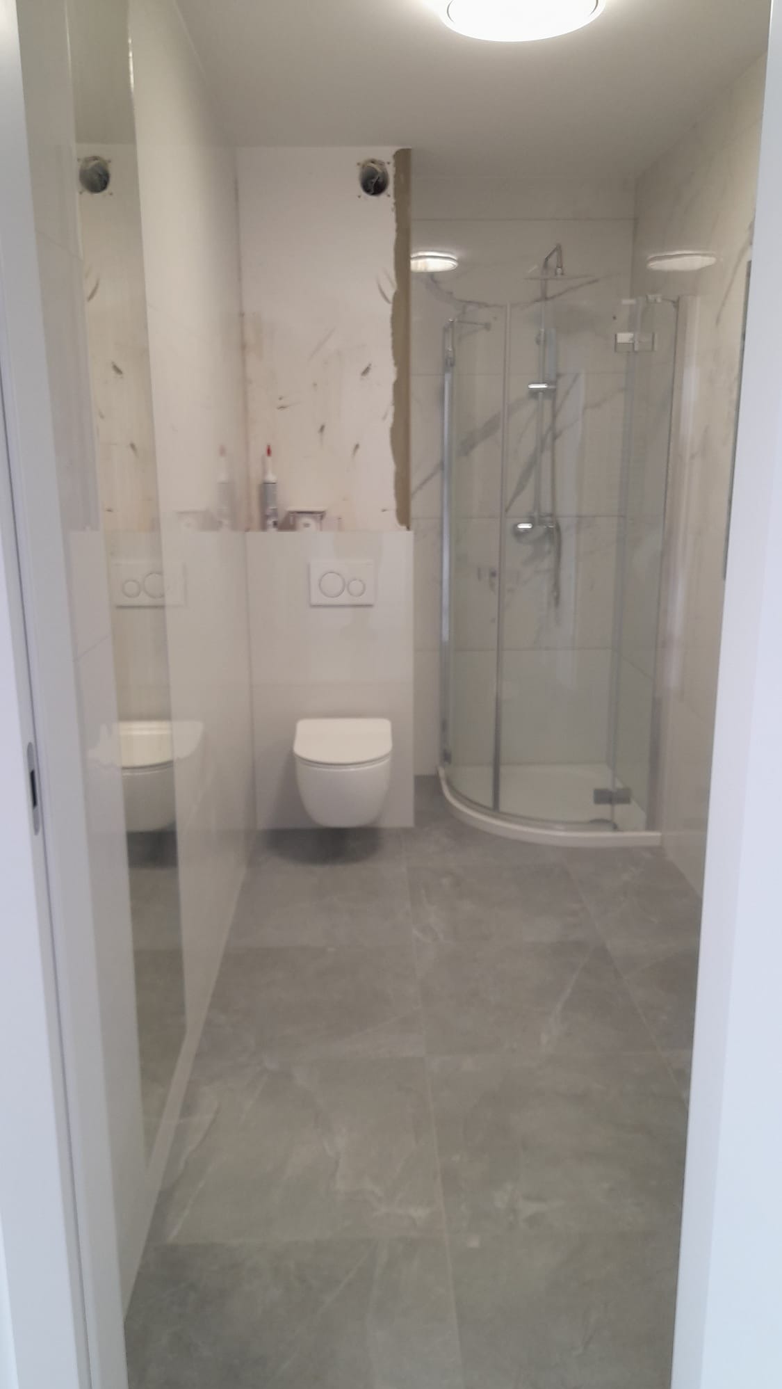 Wnętrze łazienki w trakcie wykańczania: szare płytki podłogowe, biała toaleta podwieszana, kabina prysznicowa z marmurowym wzorem, ściana częściowo przygotowana do wykończenia.