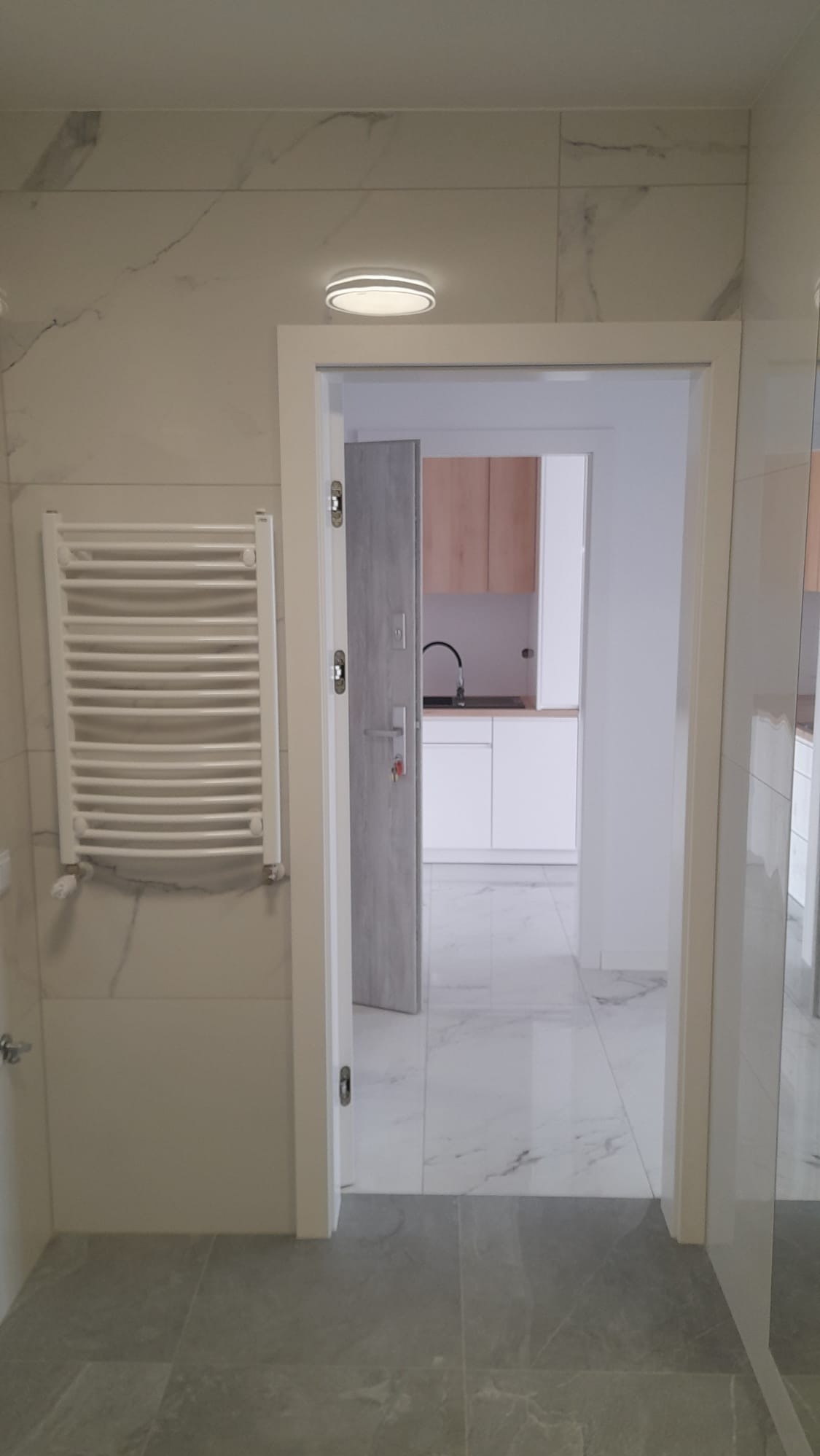 Biała łazienka z grzejnikiem drabinkowym i marmurowymi płytkami, widok na kuchnię przez otwarte drzwi. Szare płytki podłogowe, minimalistyczny design.