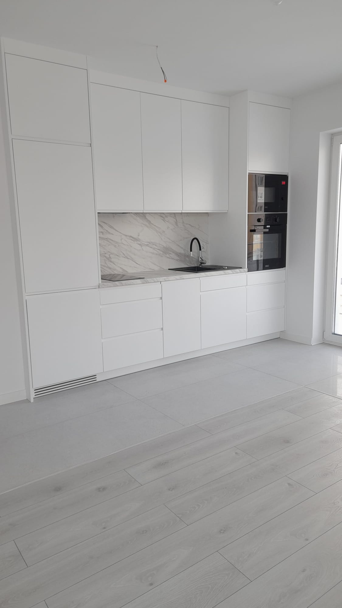 Nowoczesna, minimalistyczna kuchnia z białymi szafkami i marmurowym backsplashiem. Czarny zlew i bateria dodają kontrastu. Jasne drewniane panele podłogowe.