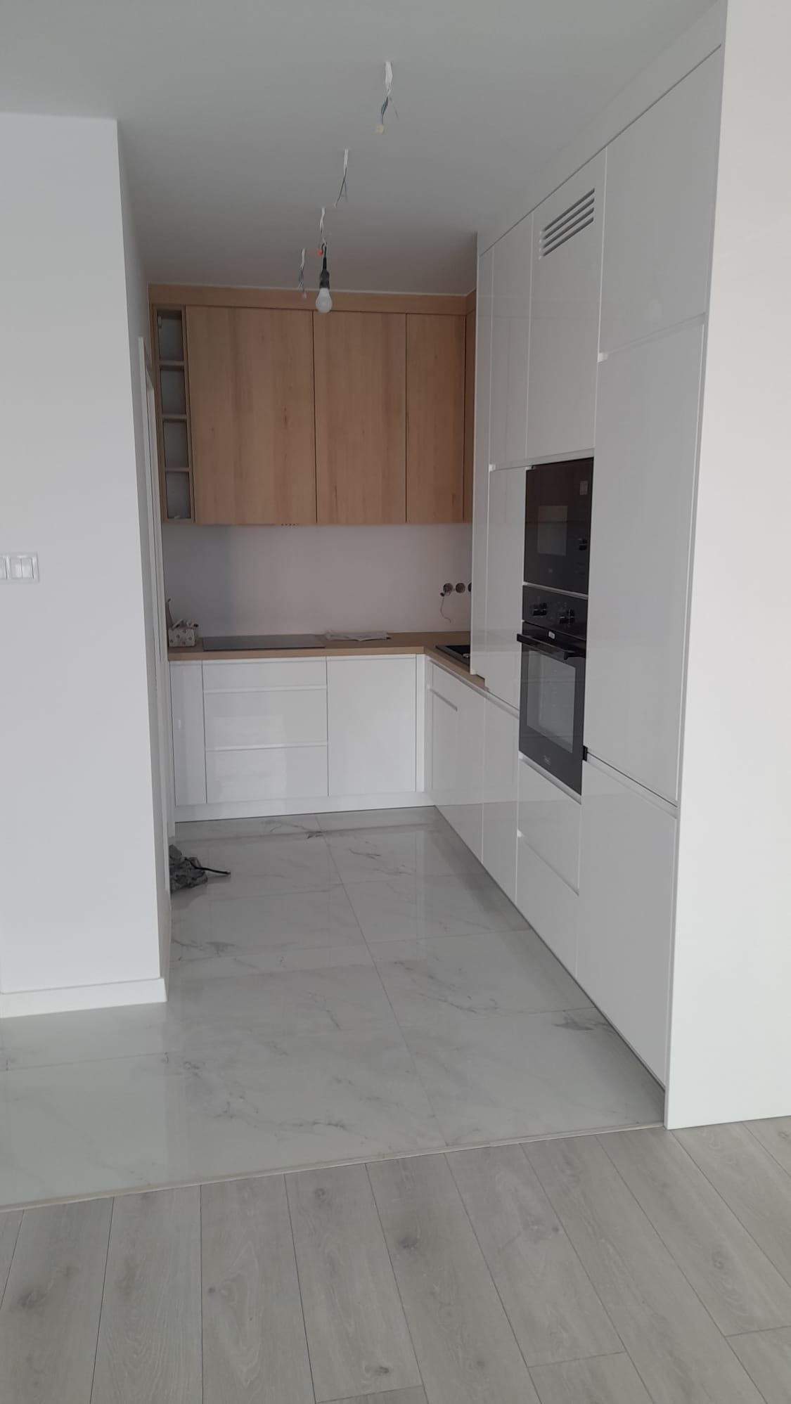 Nowoczesna kuchnia z białymi, lakierowanymi szafkami i drewnianymi górnymi szafkami. Marmurowe płytki podłogowe i widoczne przewody elektryczne.