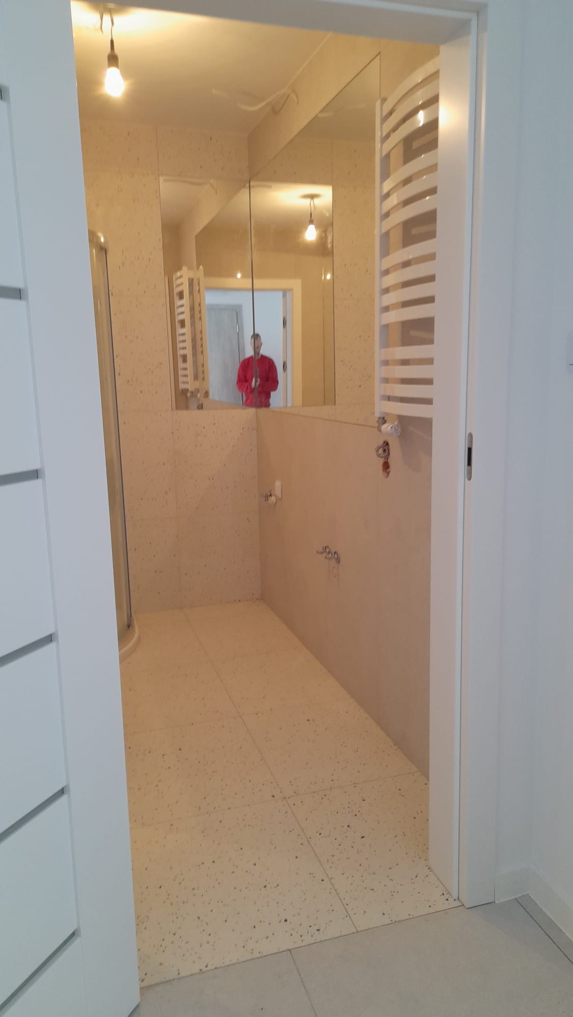 Wykończona łazienka z beżowymi płytkami lastryko na podłodze i ścianach, widoczna kabina prysznicowa i grzejnik drabinkowy. W lustrze odbija się osoba w czerwonej bluzie.