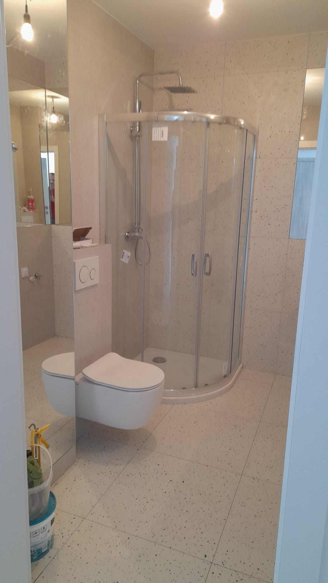 Nowoczesna łazienka po remoncie: kabina prysznicowa, podwieszana toaleta, beżowe płytki z drobnym wzorem. Widoczne odbicie w lustrze.