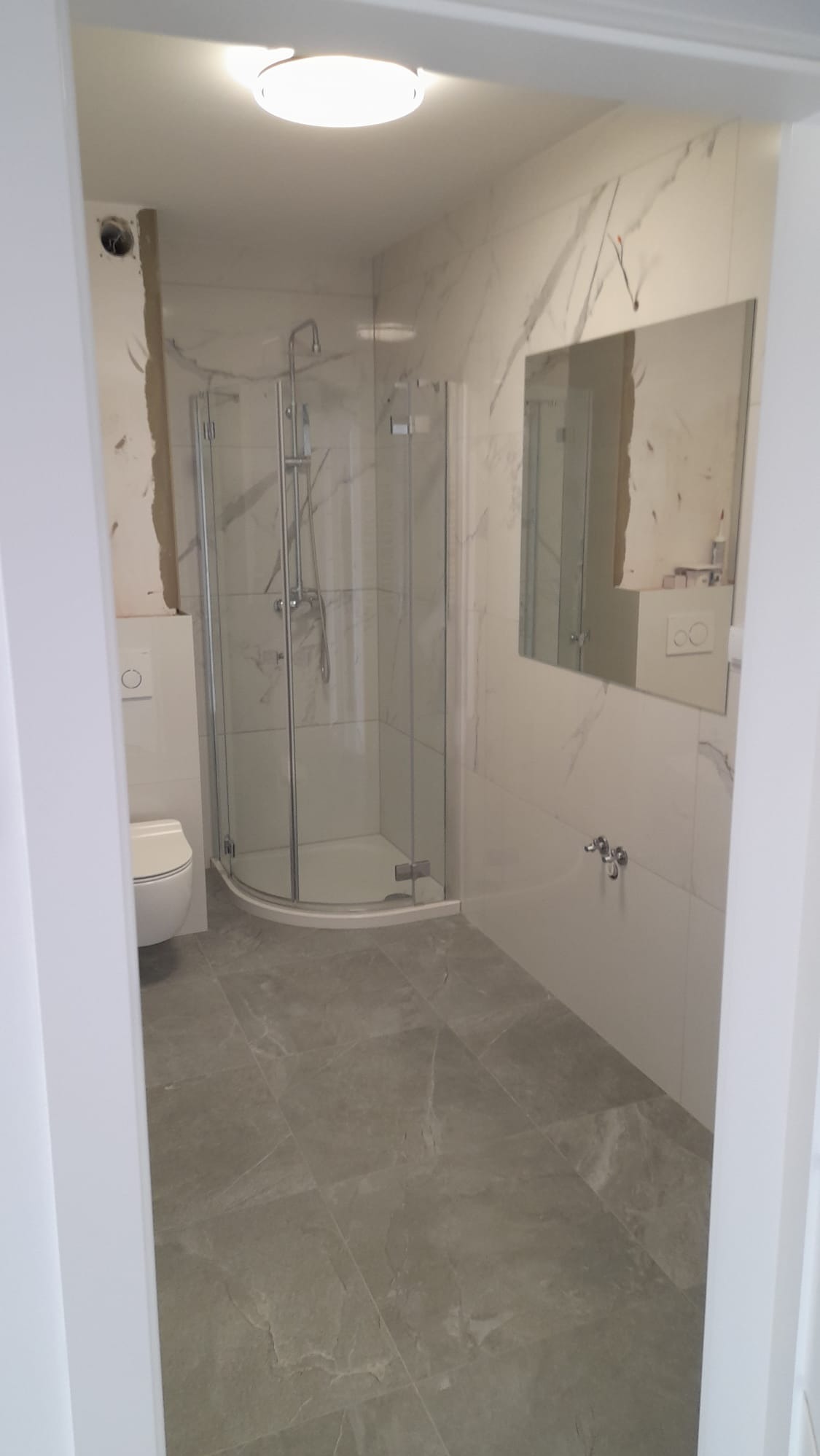 Wykończona łazienka z kabiną prysznicową typu walk-in, szarymi płytkami podłogowymi i marmurowymi płytkami ściennymi. Widok zza futryny drzwi.