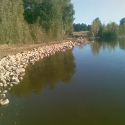 Brzeg zbiornika wodnego umocniony kamieniami, z widocznym pomostem i zielenią w tle.
