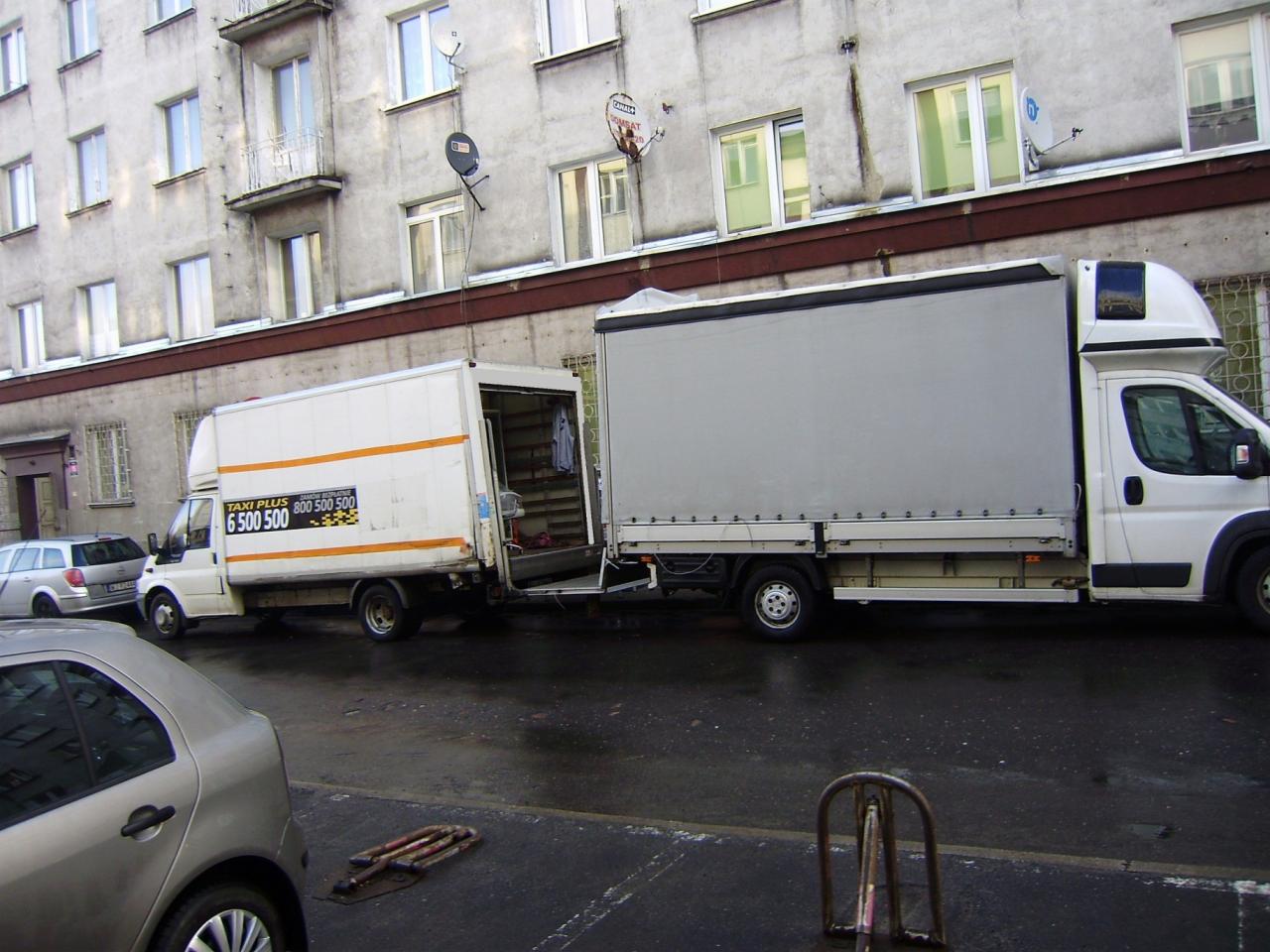 Dwa busy transportowe zaparkowane na ulicy przed budynkiem mieszkalnym; jeden z otwartymi drzwiami, drugi z plandeką, widoczne numery telefonów na boku pojazdu 'Taxi Plus 6 500 500'.