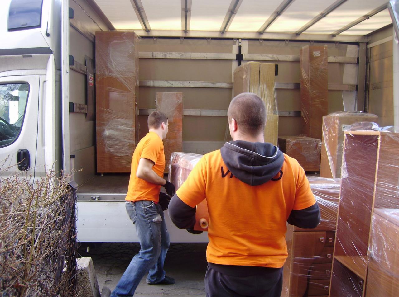 Dwóch mężczyzn w pomarańczowych koszulkach wnosi meble owinięte folią do wnętrza furgonetki, widok od tyłu, z zewnątrz widoczny fragment białego samochodu dostawczego i krzew.
