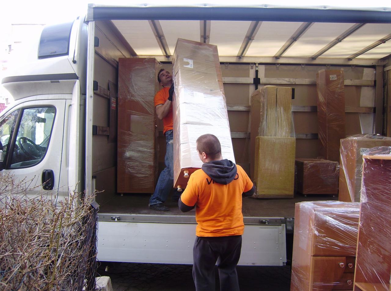 Dwóch mężczyzn w pomarańczowych koszulkach wnosi owinięty folią mebel do wnętrza furgonetki, widoczne inne zapakowane meble w przestrzeni ładunkowej.