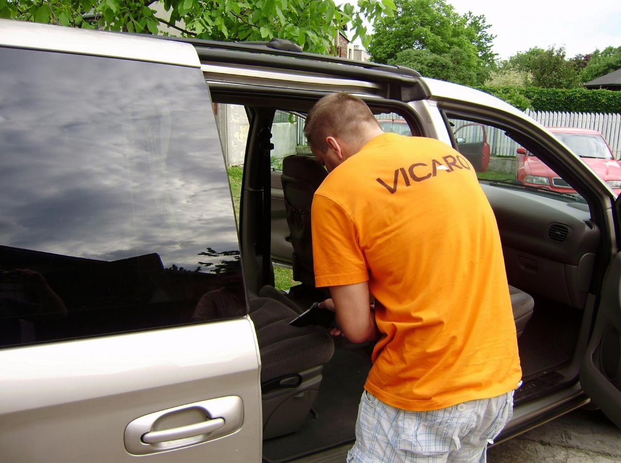 Mężczyzna w pomarańczowej koszulce z napisem VICARO odkurza wnętrze srebrnego vana z otwartymi przesuwnymi drzwiami, w tle widoczny czerwony samochód i zielona roślinność.
