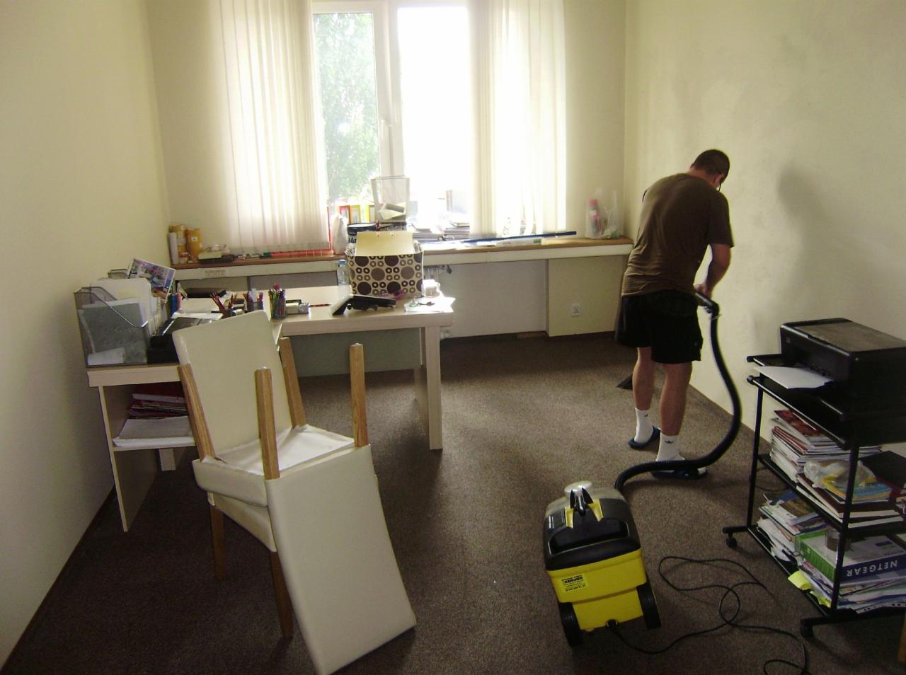 Mężczyzna odkurza brązowy dywan w biurze używając żółto-czarnego odkurzacza Karcher, z tyłu przewrócone białe krzesło i biurko z akcesoriami biurowymi oraz regał z papierami.