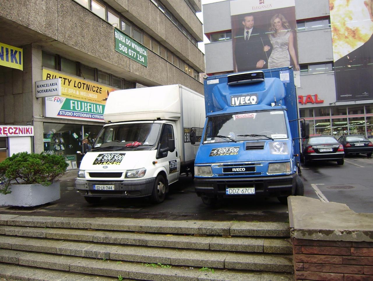 Dwa busy transportowe, biały Ford Transit i niebieski Iveco, zaparkowane przed budynkiem z szyldami reklamowymi i biurowymi, widoczne schody wejściowe.