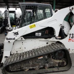 Boczny widok białej ładowarki gąsienicowej Bobcat T300 HF z czarnymi gąsienicami i logiem Bobcat, stojącej na betonowej powierzchni z innymi maszynami w tle.