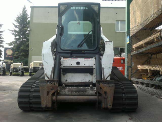 Widok z przodu na białą, lekko zabrudzoną ładowarkę gąsienicową Bobcat T300 HF z kabiną, stojącą na placu budowy. W tle budynek i reklama Shell.