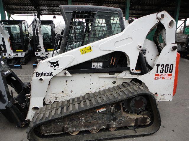 Boczny widok białej ładowarki gąsienicowej Bobcat T300 HF z czarnymi gąsienicami i logiem Bobcat, stojącej na betonowej powierzchni z innymi maszynami w tle.