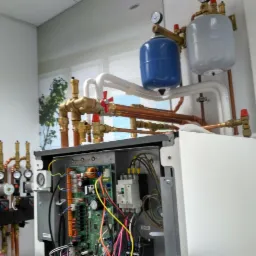 Widok z bliska na otwarty piec gazowy z widocznymi elementami instalacji, w tym miedziane rury, zawory z czerwonymi pokrętłami, manometry i zbiorniki wyrównawcze, na tle ściany z fototapetą...