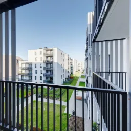 Widok z balkonu na nowoczesne osiedle z zielonym dziedzińcem, czarne metalowe barierki balkonu na pierwszym planie.
