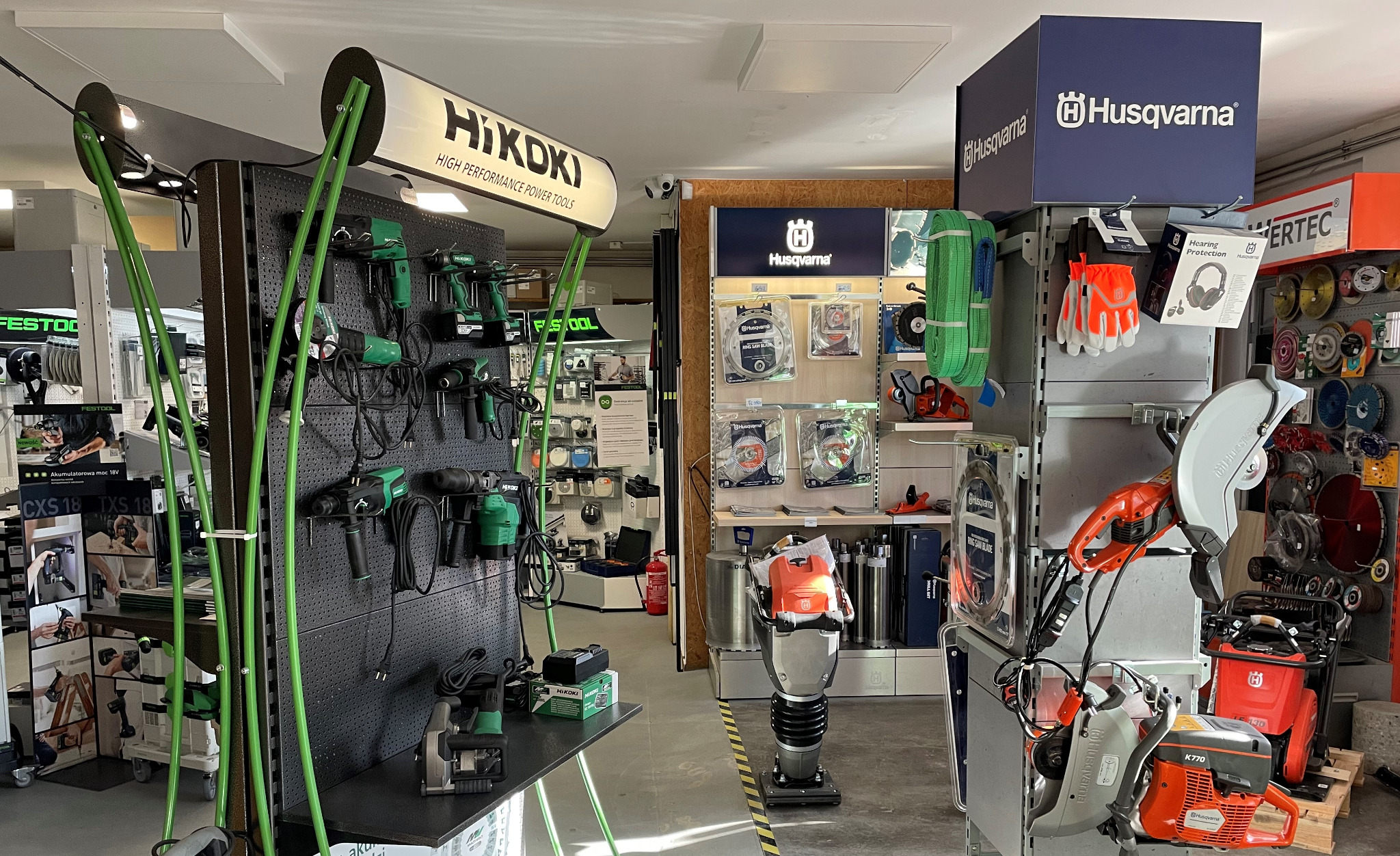 Wnętrze sklepu narzędziowego z ekspozycją elektronarzędzi marki Hikoki na perforowanej ścianie oraz sprzętu budowlanego Husqvarna, w tym ubijarki i przecinarki do betonu.