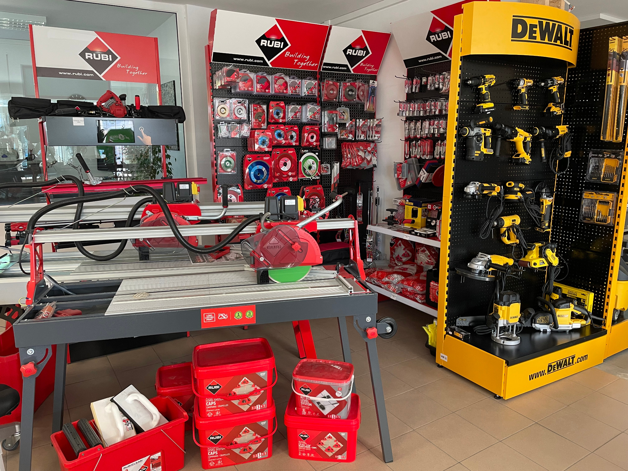 Ekspozycja w sklepie z narzędziami: przecinarki do płytek Rubi na stole z akcesoriami, obok żółty regał z elektronarzędziami DeWalt.