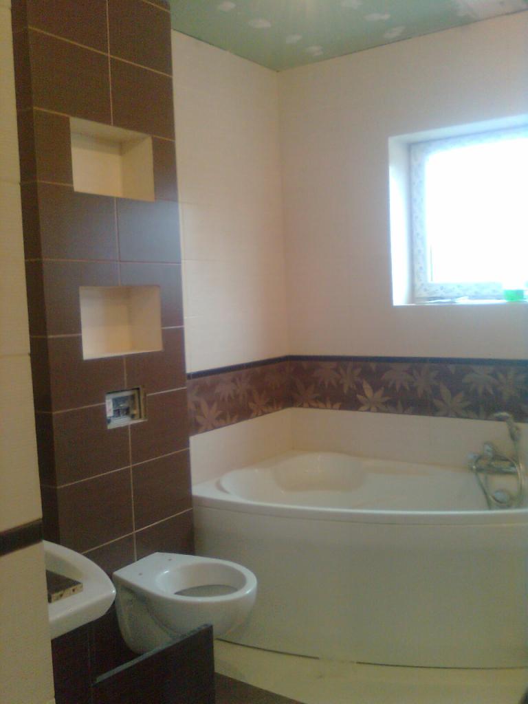 Wykończona łazienka z narożną wanną, brązowymi płytkami z wnękami i toaletą w kolorze białym, widok na okno z parapetem.