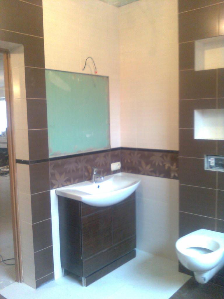 Wykończona łazienka z umywalką na szafce z ciemnego drewna, toaletą w tle i brązowymi płytkami z motywem liści na ścianie.