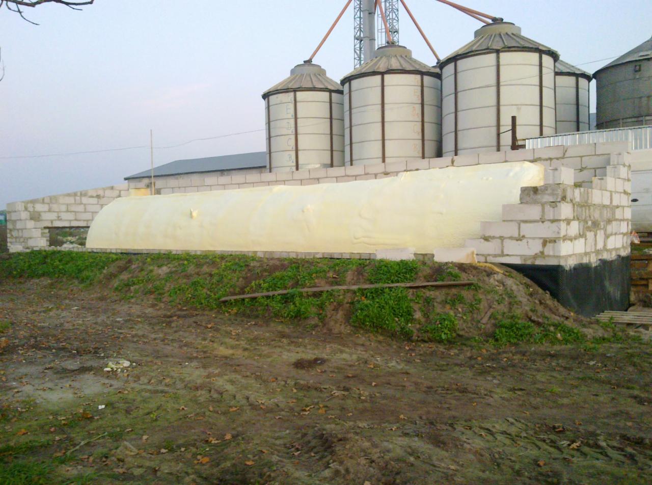 Fundament budynku gospodarczego z bloków betonowych częściowo pokryty jasną pianą izolacyjną, widoczne silosy zbożowe w tle.