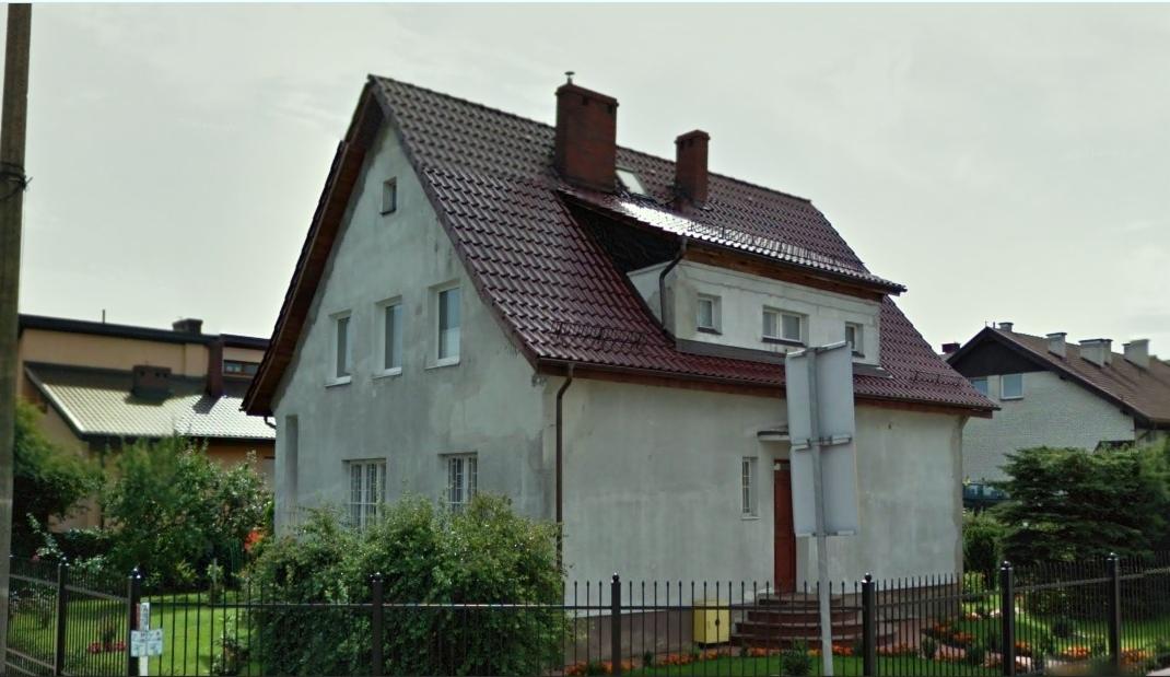 Dom z dwuspadowym dachem z czerwonej dachówki ceramicznej, widoczne kominy, rynny, elewacja w jasnym kolorze, ogrodzenie z metalowych sztachet.