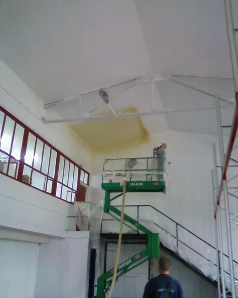 Pracownik na podnośniku maluje ścianę wewnątrz hali, widoczne okna z czerwonymi ramami i elementy konstrukcyjne dachu.