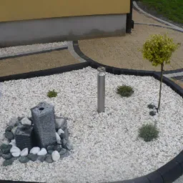 Ogród z dekoracyjną fontanną z kamienia, białymi kamykami, metalowym słupkiem oświetleniowym i ozdobnym drzewkiem na tle kostki brukowej w odcieniach beżu i czerni.