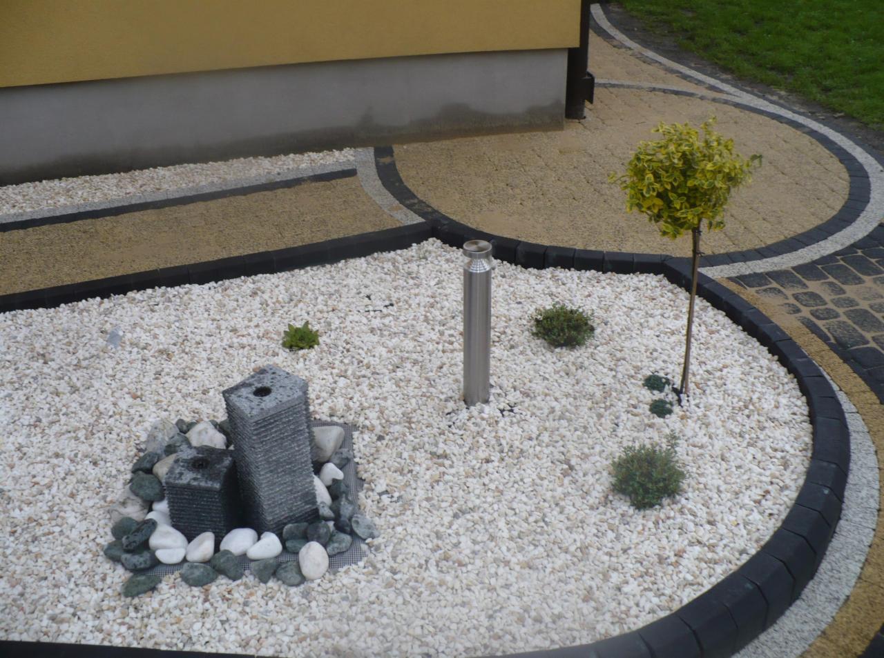 Ogród z dekoracyjną fontanną z kamienia, białymi kamykami, metalowym słupkiem oświetleniowym i ozdobnym drzewkiem na tle kostki brukowej w odcieniach beżu i czerni.