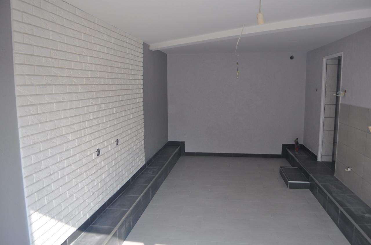 Wnętrze pomieszczenia po pracach wykończeniowych w Gaworzycach: biała ściana z imitacją cegły, szara podłoga z ciemnymi listwami, surowe szare ściany, widoczny fragment instalacji elektrycznej.