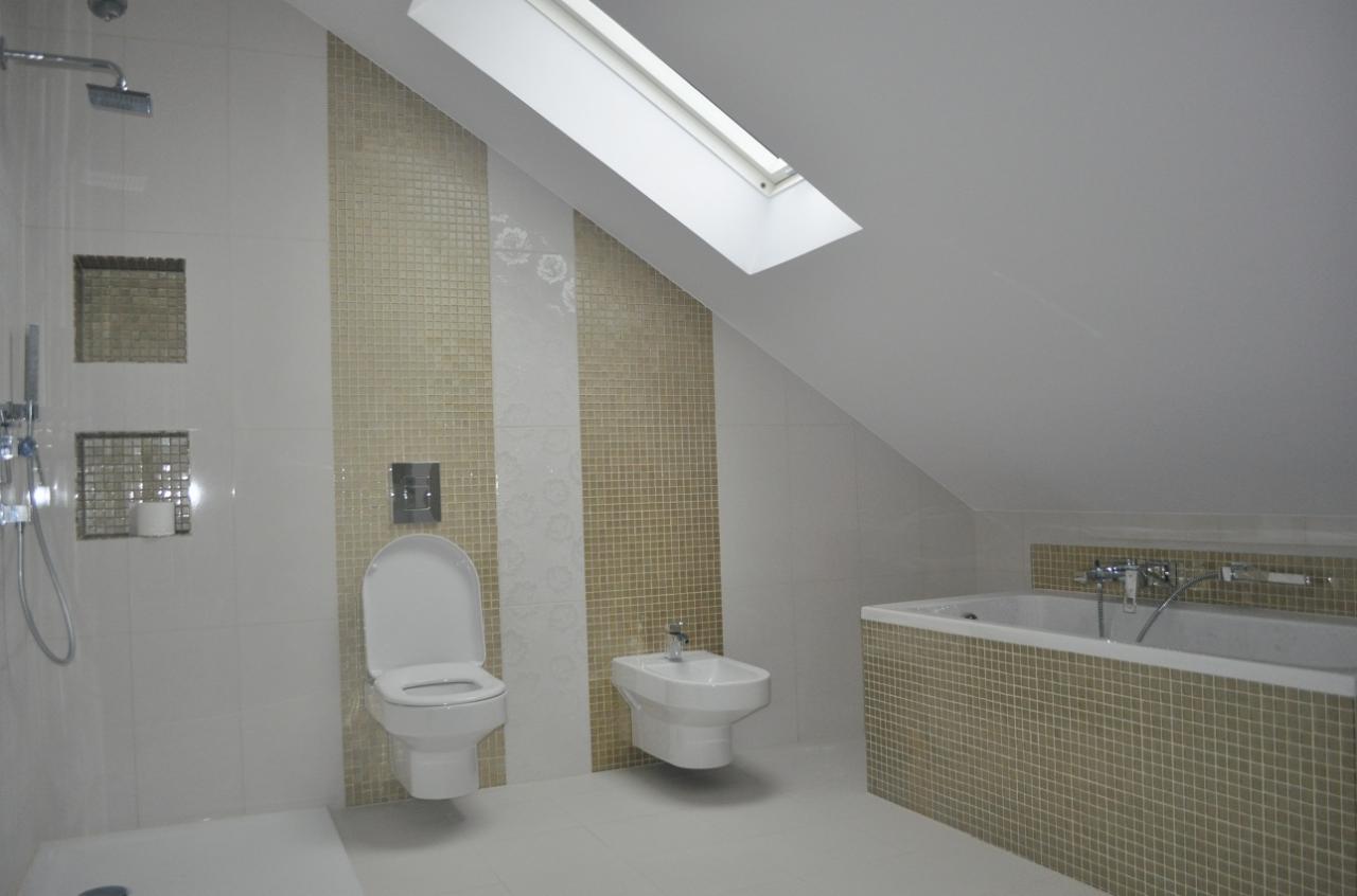 Wykończona łazienka na poddaszu z białą armaturą, mozaiką w odcieniach beżu i dużym oknem dachowym, dająca wrażenie przestronności i jasności.