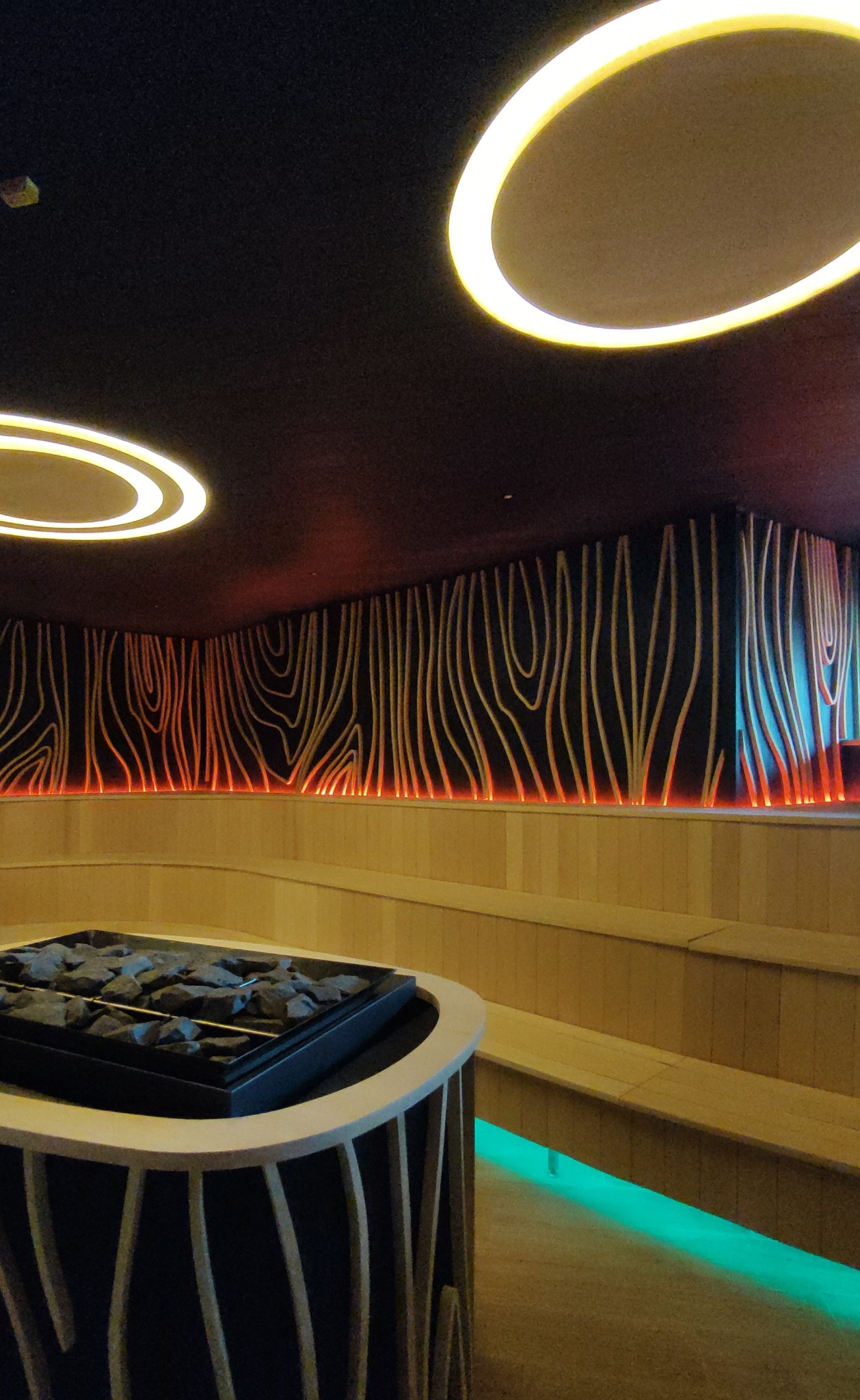 Nowoczesna sauna z dekoracyjnym oświetleniem LED: okrągłe lampy sufitowe, podświetlana ściana z motywem drewna i zielone podświetlenie ławek.