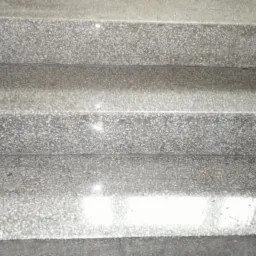 Trzy stopnie schodów wykończone szarym kamieniem z drobnym wzorem, widoczne z góry pod kątem. Jasne odbicie światła na powierzchni.