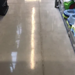 Szara, gładka podłoga z widocznym dylatacją pośrodku, odbijająca światło, z fragmentami regałów sklepowych po bokach.