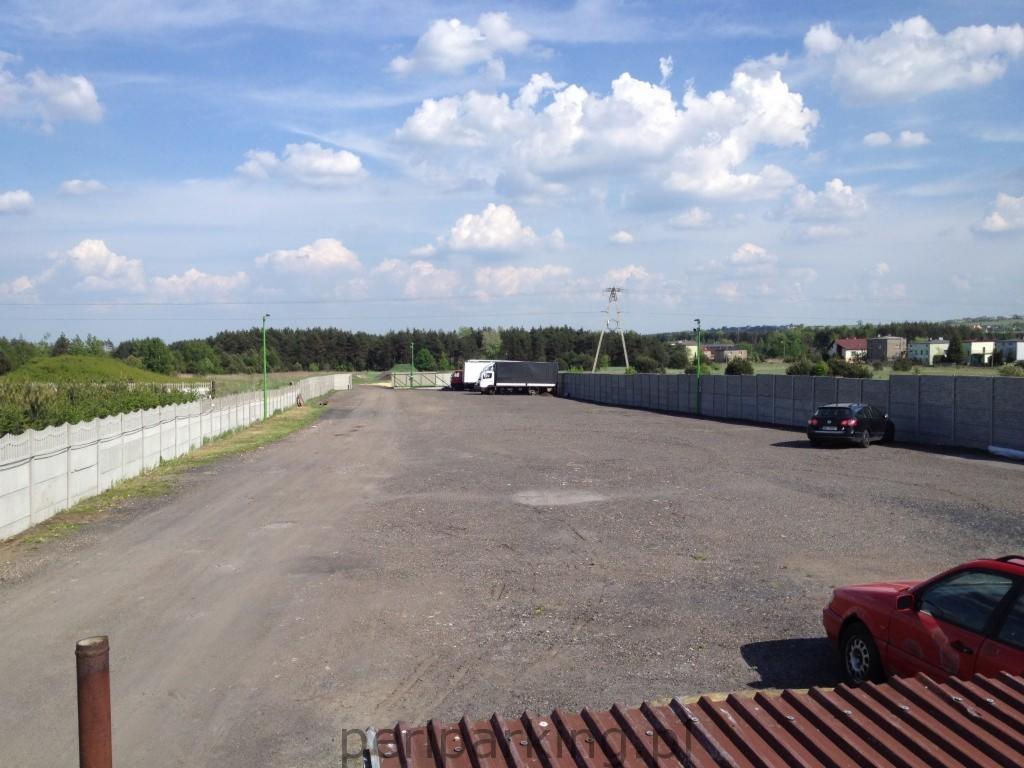 Rozległy, szutrowy parking z zaparkowanymi samochodami osobowymi i ciężarowym, otoczony betonowym ogrodzeniem, w tle widoczny las i zabudowania.