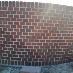 Ceglany, zakrzywiony mur ogrodzeniowy w trakcie budowy, widoczne niedociągnięcia w podłożu i wystające cegły na górze.