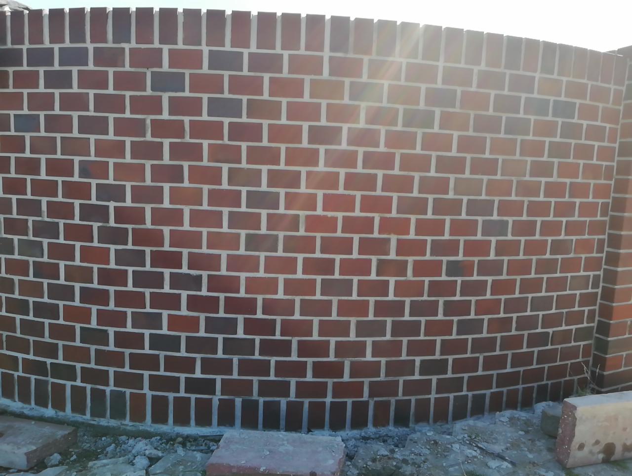 Ceglany, zakrzywiony mur ogrodzeniowy w trakcie budowy, widoczne niedociągnięcia w podłożu i wystające cegły na górze.