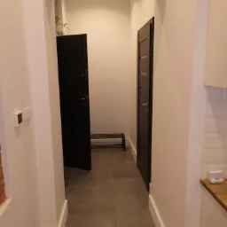 Wnętrze przedpokoju z dwoma czarnymi drzwiami, szarą podłogą z płytek i minimalistyczną ławką na buty, widok z perspektywy wnętrza mieszkania, z widoczną instalacją elektryczną pod sufitem.