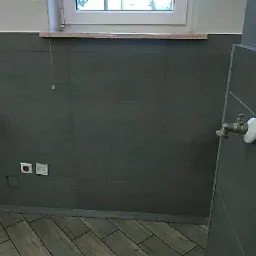 Szare płytki ścienne i podłogowe w łazience w Suszu, widoczny kran na ścianie oraz gniazdka elektryczne.