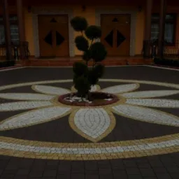 Ozdobny podjazd z kostki brukowej ułożonej w motyw kwiatowy, z drzewkiem w centrum. Ciemna kostka obramowana jaśniejszymi elementami, budynek w tle.