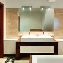 Nowoczesna łazienka z umywalką nablatową na ciemnobrązowej szafce, duże lustro z trzema punktami świetlnymi, jasne ściany z elementami dekoracyjnymi w odcieniach beżu i brązu, drewniane drzwi po...