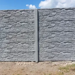 Gotowy, szary, betonowy płot imitujący kamień, z widocznymi słupkami, na tle nieba z chmurami.
