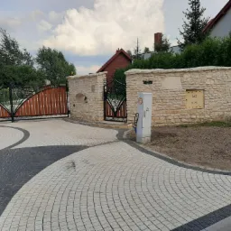 Nowoczesny podjazd z szarej i białej kostki brukowej w łukowaty wz&oacute;r prowadzący do drewnianej bramy wjazdowej z metalowymi zdobieniami i kamiennego ogrodzenia z małą furtką z czarnymi metalowymi...