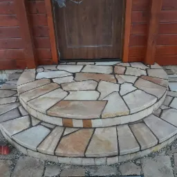 Dwu-stopniowe schody wejściowe do domu, wyłożone nieregularnymi płytami kamiennymi w odcieniach beżu i brązu, z ciemnymi fugami, prowadzące do drewnianych drzwi i elewacji z drewnianych desek.