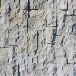 Ściana wyłożona nieregularnymi, jasnymi płytkami z kamienia naturalnego o chropowatej fakturze, ułożonymi bez fugi, w ciepłym świetle słonecznym.