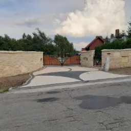 Wjazd na posesję z drewnianą bramą z metalowymi zdobieniami, otoczony niskim murem z jasnego kamienia i wybrukowaną nawierzchnią w odcieniach szarości i bieli.