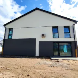 Nowoczesny dom jednorodzinny z biało-grafitową elewacją, widoczne okna z czarnymi ramami, zamontowany klimatyzator na ścianie, teren wokół domu w trakcie prac ziemnych.