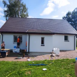 Prace wykończeniowe elewacji domu jednorodzinnego: malowanie ściany zewnętrznej na biało, pracownik na podeście, widoczne narzędzia i materiały budowlane, trawnik w pierwszym planie.